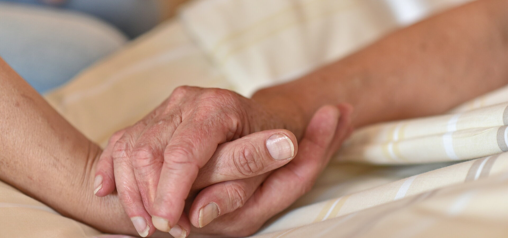 Zwei Hände halten sich sanft fest. Eine menschliche Hand mit hellen Nägeln greift nach einer älteren Hand mit einem warmen und gefühlvollen Ausdruck. Im Hintergrund ist ein verschwommener Teil einer anderen Person sichtbar. Die Atmosphäre wirkt intim und beschützend.