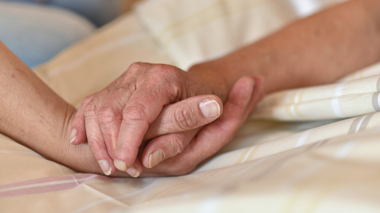 Zwei Hände halten sich sanft fest. Eine menschliche Hand mit hellen Nägeln greift nach einer älteren Hand mit einem warmen und gefühlvollen Ausdruck. Im Hintergrund ist ein verschwommener Teil einer anderen Person sichtbar. Die Atmosphäre wirkt intim und beschützend.