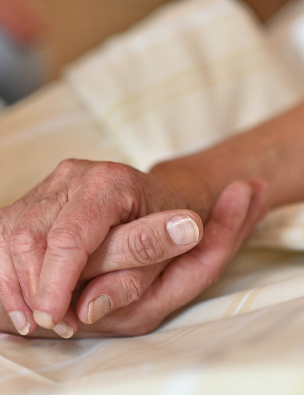 Zwei Hände halten sich sanft fest. Eine menschliche Hand mit hellen Nägeln greift nach einer älteren Hand mit einem warmen und gefühlvollen Ausdruck. Im Hintergrund ist ein verschwommener Teil einer anderen Person sichtbar. Die Atmosphäre wirkt intim und beschützend.