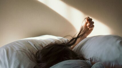 Eine Person liegt mit dem Rücken auf einem Bett, das Gesicht verborgen. Ihr Arm ist nach hinten gestreckt und greift nach den Haaren. Sanftes Licht fällt von einer Wand auf die Bettdecke, wodurch eine ruhige und entspannende Atmosphäre entsteht.