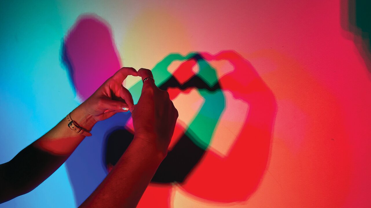 Zwei Hände formen ein Herzsymbol, das als Schatten auf einer bunten Wand zu sehen ist. Die Schatten sind in verschiedenen Farben wie Rot, Blau und Grün.