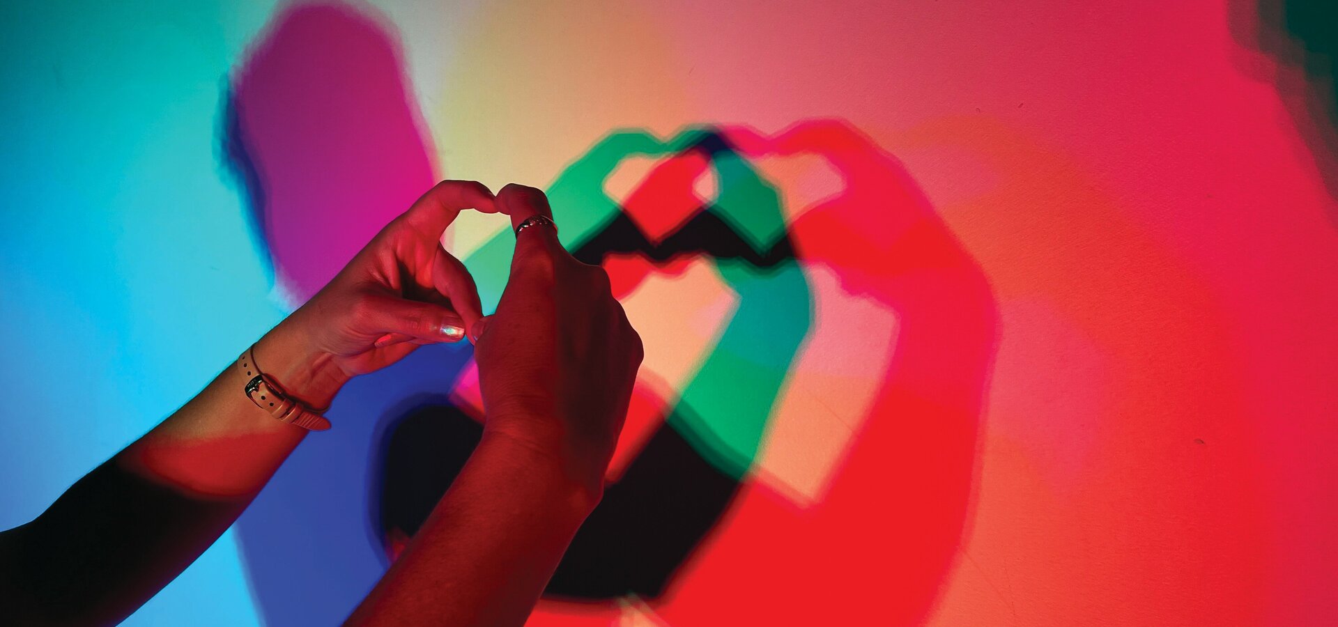 Zwei Hände formen ein Herzsymbol, das als Schatten auf einer bunten Wand zu sehen ist. Die Schatten sind in verschiedenen Farben wie Rot, Blau und Grün.