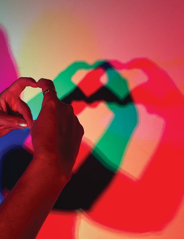 Zwei Hände formen ein Herzsymbol, das als Schatten auf einer bunten Wand zu sehen ist. Die Schatten sind in verschiedenen Farben wie Rot, Blau und Grün.