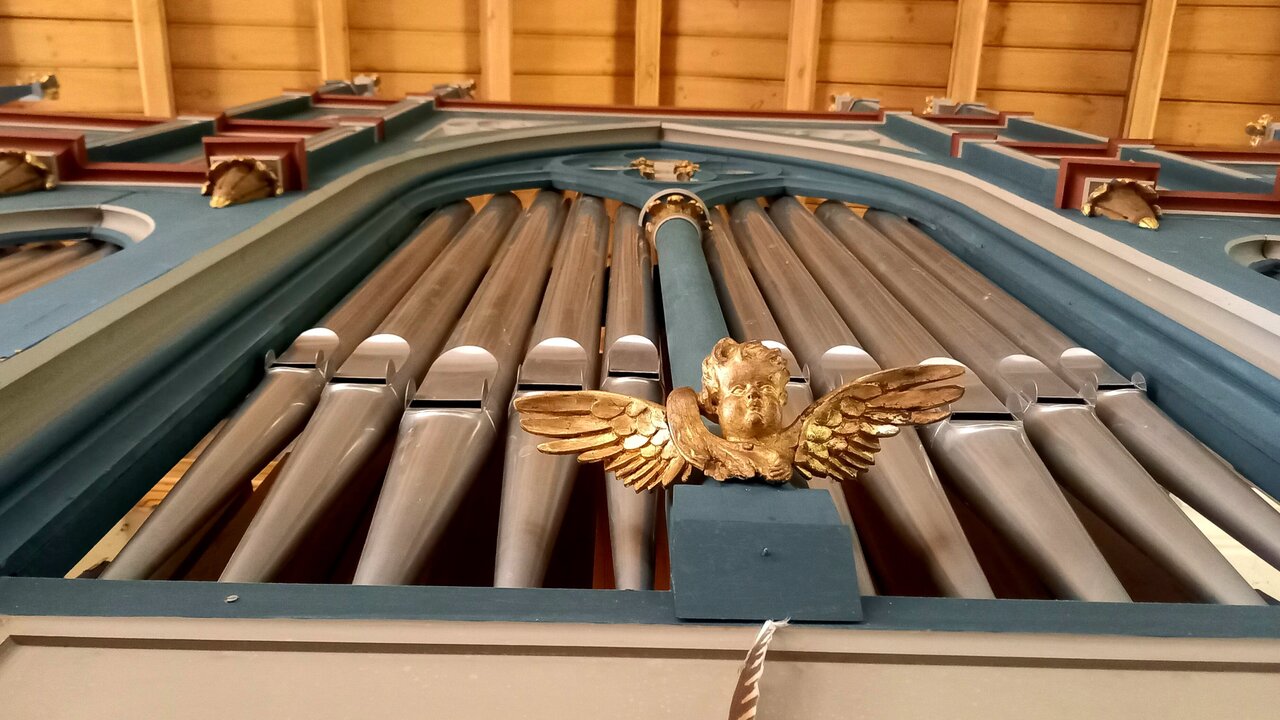 Eine prunkvolle Kirchenorgel, die von unten fotografiert wurde. Die Pfeifen sind in mehreren Reihen angeordnet, während oben ein kunstvolles, goldenes Ornament mit Flügeln zu sehen ist. Die Decke ist aus Holz und trägt zur feierlichen Atmosphäre bei.