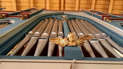 Eine prunkvolle Kirchenorgel, die von unten fotografiert wurde. Die Pfeifen sind in mehreren Reihen angeordnet, während oben ein kunstvolles, goldenes Ornament mit Flügeln zu sehen ist. Die Decke ist aus Holz und trägt zur feierlichen Atmosphäre bei.