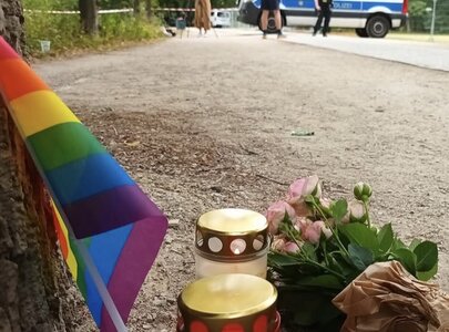 Am Boden liegen zwei Kerzen mit der Aufschrift „R.I.P.“ und rosa Blumen. Im Hintergrund steht ein Polizeiwagen, während Beamte im Bereich ermitteln. Eine Regenbogenfahne weht in der Nähe, was auf eine Gedenkfeier für eine Person hindeutet.