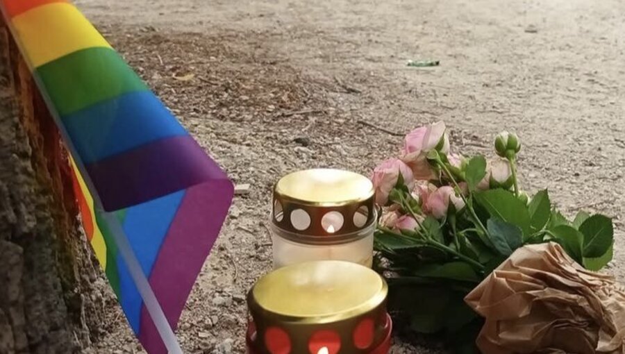 Am Boden liegen zwei Kerzen mit der Aufschrift „R.I.P.“ und rosa Blumen. Im Hintergrund steht ein Polizeiwagen, während Beamte im Bereich ermitteln. Eine Regenbogenfahne weht in der Nähe, was auf eine Gedenkfeier für eine Person hindeutet.
