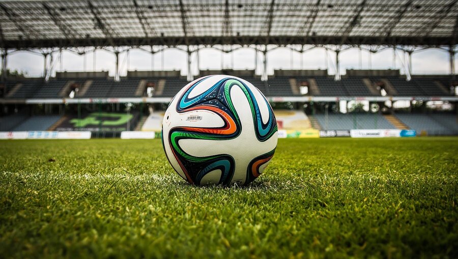 Ein Fußball liegt auf dem grünen Rasen eines Stadions. Im Hintergrund sind Sitzreihen und die Struktur des Daches erkennbar, während der Platz für das Spiel vorbereitet ist. Die Atmosphäre ist sportlich und lebendig.