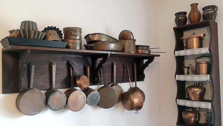 Eine Holzregalwand zeigt verschiedene Kupferküchenutensilien, darunter Töpfe, Pfannen und Backformen. An einer Seitenwand sind weitere Kupfergefäße in Regalen angeordnet. Die Anordnung vermittelt ein nostalgisches Küchendesign und erinnert an traditionelle Kochmethoden.