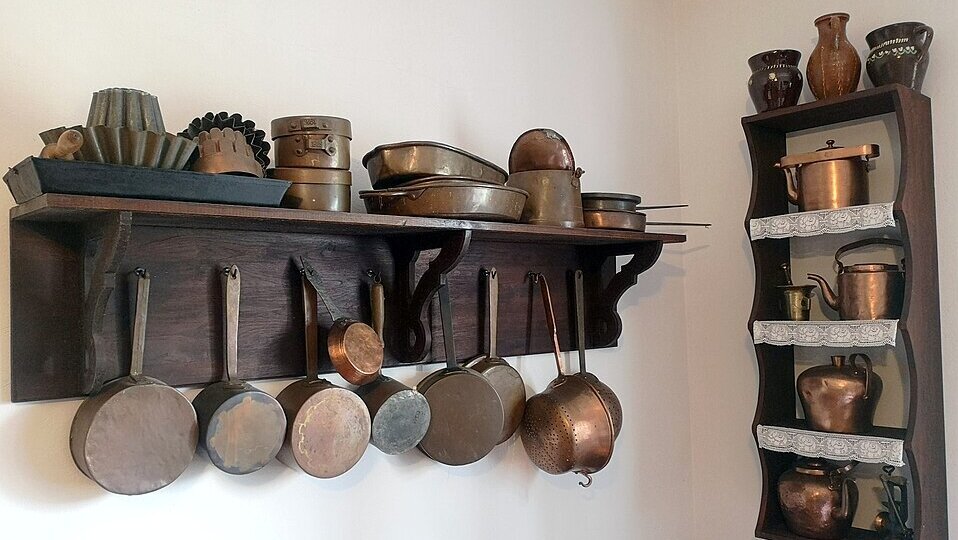 Eine Holzregalwand zeigt verschiedene Kupferküchenutensilien, darunter Töpfe, Pfannen und Backformen. An einer Seitenwand sind weitere Kupfergefäße in Regalen angeordnet. Die Anordnung vermittelt ein nostalgisches Küchendesign und erinnert an traditionelle Kochmethoden.