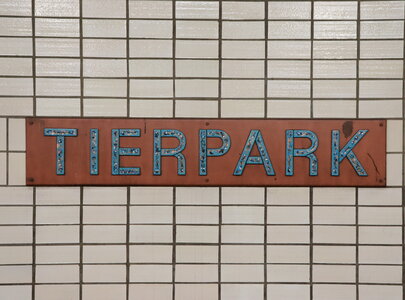 Ein rostiger Schild mit der Aufschrift "TIERPARK" in blauen Buchstaben, angebracht an einer wand aus weißen Fliesen.