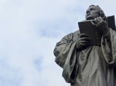 Die Statue eines Mannes, der ein Buch in der Hand hält, steht aufrecht. Er trägt einen langen Mantel und schaut nach oben. Der Hintergrund zeigt einen hellen Himmel mit wenigen Wolken.