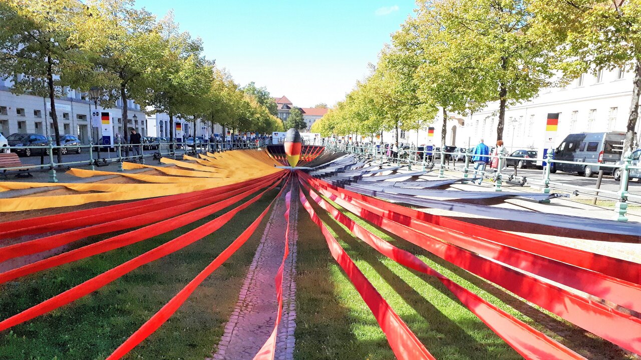 Ein weitläufiger Platz mit einer Wiese in der Mitte, umgeben von Bäumen. In der Mitte verläuft ein schmaler, geometrischer Weg, der von roten Bändern flankiert wird. Diese Bänder ziehen sich in die Entfernung und schaffen eine dynamische Perspektive. Die Szenerie vermittelt eine moderne, künstlerische Atmosphäre.
