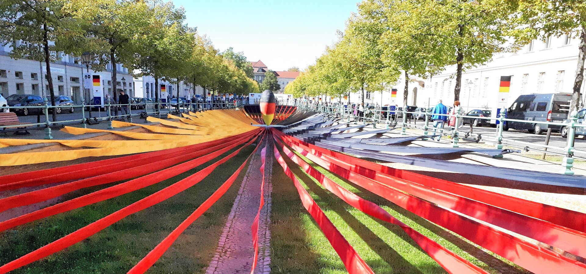 Ein weitläufiger Platz mit einer Wiese in der Mitte, umgeben von Bäumen. In der Mitte verläuft ein schmaler, geometrischer Weg, der von roten Bändern flankiert wird. Diese Bänder ziehen sich in die Entfernung und schaffen eine dynamische Perspektive. Die Szenerie vermittelt eine moderne, künstlerische Atmosphäre.