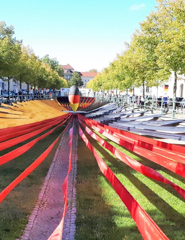 Ein weitläufiger Platz mit einer Wiese in der Mitte, umgeben von Bäumen. In der Mitte verläuft ein schmaler, geometrischer Weg, der von roten Bändern flankiert wird. Diese Bänder ziehen sich in die Entfernung und schaffen eine dynamische Perspektive. Die Szenerie vermittelt eine moderne, künstlerische Atmosphäre.