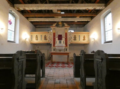 Das Bild zeigt den Innenraum einer kleinen Kirche mit einem schlichten Altar im Zentrum. Der Altar ist farbenfroh mit Schmuckelementen verziert, umgeben von Holzbänken für die Gemeinde. Sanftes Licht fällt durch die Fenster und beleuchtet die historischen Elemente der Kirche.