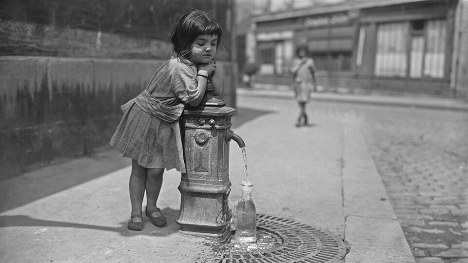 Ein kleines Mädchen lehnt an einem Wasserbrunnen, während Wasser in eine Flasche fließt. Im Hintergrund ist ein weiteres Kind zu sehen, das die Straße entlanggeht. Die Szenerie vermittelt einen Eindruck von Vergangenheit und Alltag.