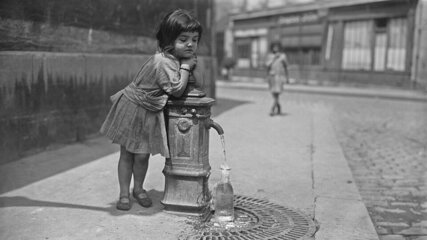Ein kleines Mädchen lehnt an einem Wasserbrunnen, während Wasser in eine Flasche fließt. Im Hintergrund ist ein weiteres Kind zu sehen, das die Straße entlanggeht. Die Szenerie vermittelt einen Eindruck von Vergangenheit und Alltag.