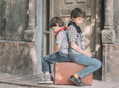 Zwei Jungen sitzen Rücken an Rücken auf einem Koffer. Der Junge links trägt einen roten Schal und schaut nach unten, während der Junge rechts einen Schal um den Hals hat und in die Ferne blickt. Die Szene vermittelt eine Stimmung von Freundschaft und Nachdenklichkeit.