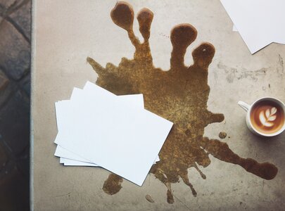 Ein Tisch mit einem Kaffeetassenabdruck und einer braunen Kaffeefleckenformation. Darauf liegen mehrere weiße Blätter, die durch die Kaffeeflecken unordentlich erscheinen. Der Kaffee in der Tasse hat ein kunstvolles Muster auf der Oberfläche.