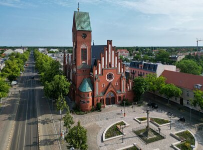 Die Aufnahme zeigt eine imposante, rotziegelne Kirche mit einem hohen Turm und markanten architektonischen Details. Umgeben von Bäumen, verläuft eine Straße vor dem Gebäude. Im Vordergrund ist ein Platz mit einem Brunnen und Sitzgelegenheiten sichtbar.