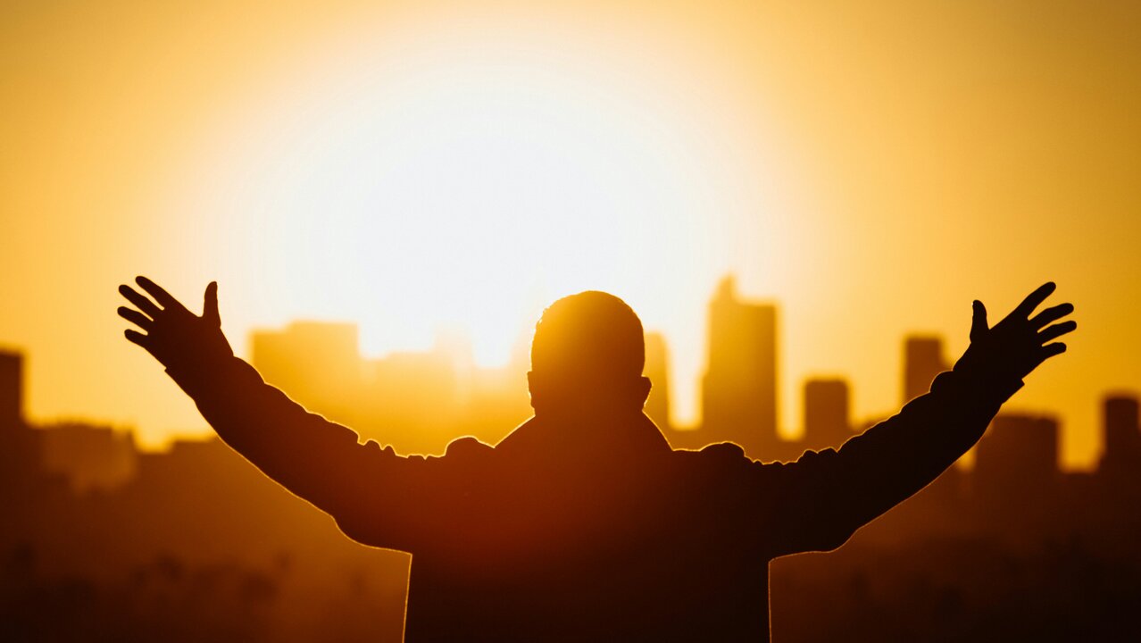 Eine silhouette einer Person mit weit ausgestreckten Armen vor einem Sonnenuntergang. Im Hintergrund ist eine Stadtlandschaft zu sehen, die von der goldenen Farbe des Sonnenlichts erleuchtet wird. Das Bild vermittelt ein Gefühl von Freiheit und Hoffnung.