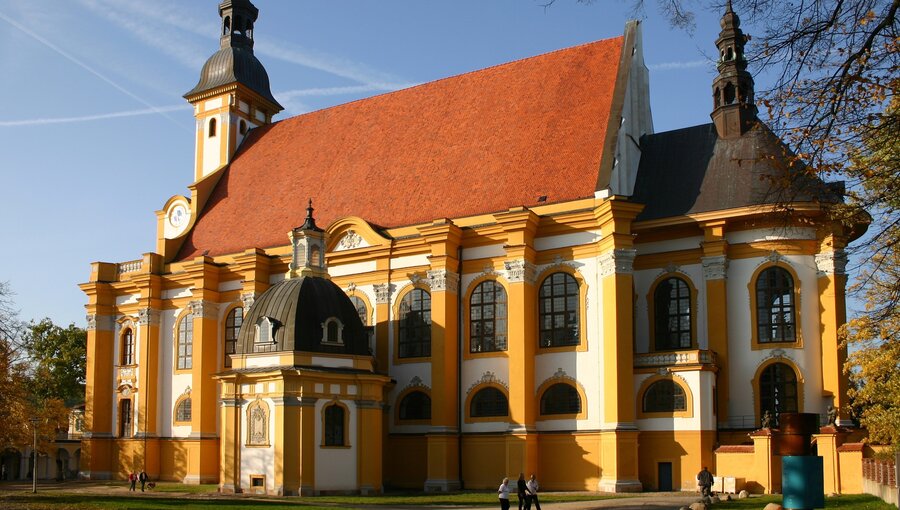 Eine große barocke Kirche mit einer markanten orangefarbenen Fassade, einem hohen Glockenturm und einem neugotischen Anbau. Beleuchtet von Sonnenlicht, steht die Kirche umgeben von Bäumen und einem klaren Himmel. Spaziergänger sind im Vordergrund zu sehen.