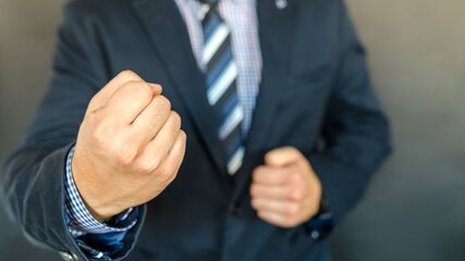 Ein Mann in einem Anzug steht mit geballter Faust da. Seine Haltung deutet auf Entschlossenheit oder Aggression hin. Der Hintergrund ist neutral und verstärkt den Fokus auf die Person und die Faust.