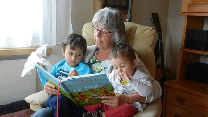 Eine Großmutter sitzt in einem Sessel und liest zwei kleinen Kindern ein Buch vor. Die Kinder sitzen auf ihren Knien, interessiert und aufmerksam. Im Hintergrund ist eine helle, freundliche Wohnung zu sehen.