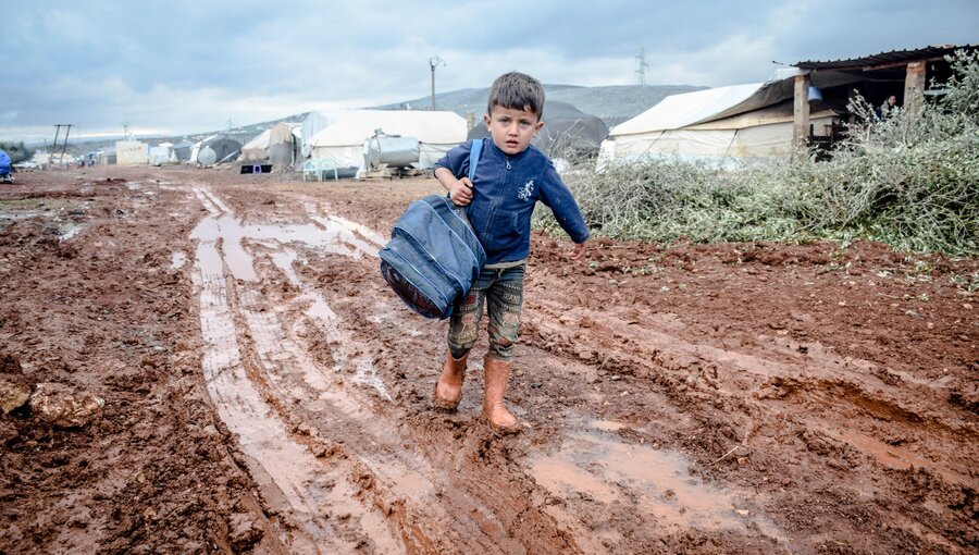 Ein kleiner Junge mit einem Rucksack geht barfuß durch einen schlammigen Weg in einem Flüchtlingslager. Das Wetter ist wolkig und die Umgebung besteht aus Zelten und unebenem, matschigem Boden.