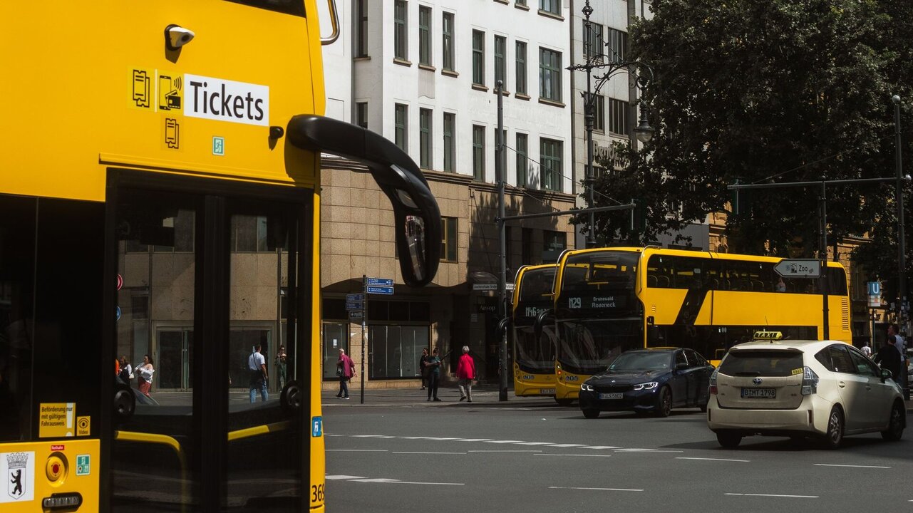 Eine gelbe Doppeldeckerbus-Haltestelle mit einem Schild für "Tickets" im Vordergrund, während im Hintergrund ein weiterer gelber Bus und mehrere Autos an einer belebten Straße anhalten. Geprägte städtische Umgebung mit Gebäuden und Bäumen.