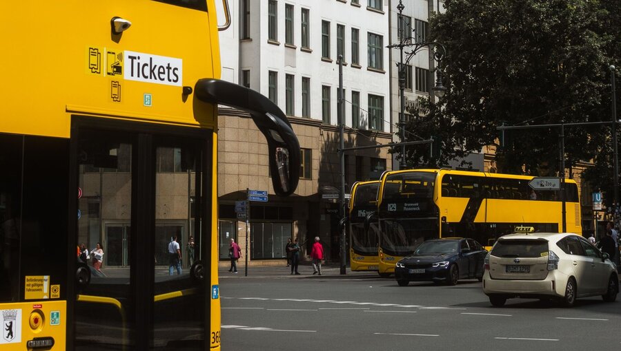 Eine gelbe Doppeldeckerbus-Haltestelle mit einem Schild für "Tickets" im Vordergrund, während im Hintergrund ein weiterer gelber Bus und mehrere Autos an einer belebten Straße anhalten. Geprägte städtische Umgebung mit Gebäuden und Bäumen.