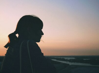 Eine Silhouette einer nachdenklichen Person, die gegen den Sonnenuntergang blickt. Im Hintergrund sind sanfte Farben des Himmels und ein Fluss sichtbar. Die Atmosphäre vermittelt Ruhe und Reflexion.