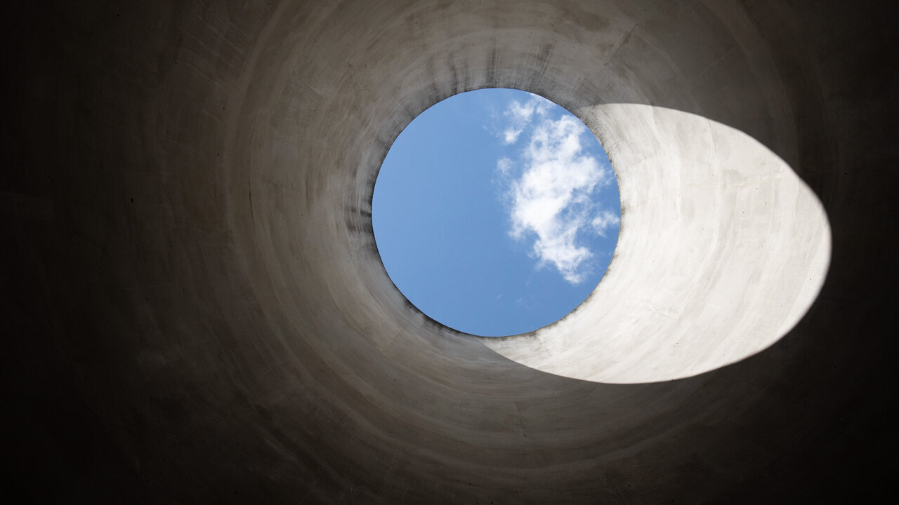 Blick durch ein großes, rundes Rohr nach oben, wo blauer Himmel und einige Wolken sichtbar sind. Der innere Rand des Rohrs ist aus grauem Beton. Hoffnung.