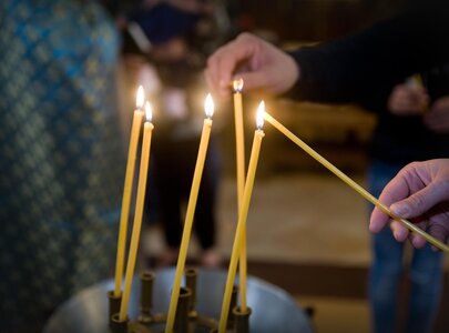 Eine Hand zündet eine Kerze an, während vier bereits brennende Kerzen in einem halbrunden Kerzenständer stehen. Die Umgebung wirkt feierlich und ruhig.