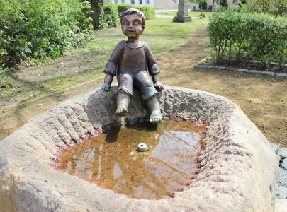 Eine bronzene Statue eines sitzenden Jungen mit lächelndem Gesicht, der mit ausgeziehter Hose an einem Steintrog sitzt. Der Trog enthält trübes Wasser und ist von einer grünen Umgebung mit Büschen umgeben.