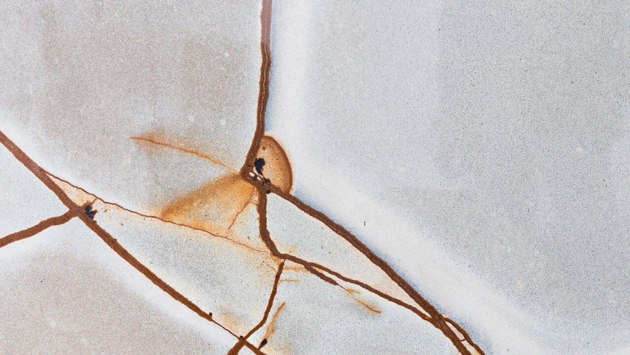 Ein abstraktes Bild einer hellen, glatten Oberfläche, auf der feine, braune Risse und Adern verlaufen. Die Risse sind unregelmäßig und verleihen der Fläche eine interessante Textur und Tiefe. Die Farbkontraste zwischen dem Hellgrau und dem Braun sind deutlich sichtbar.