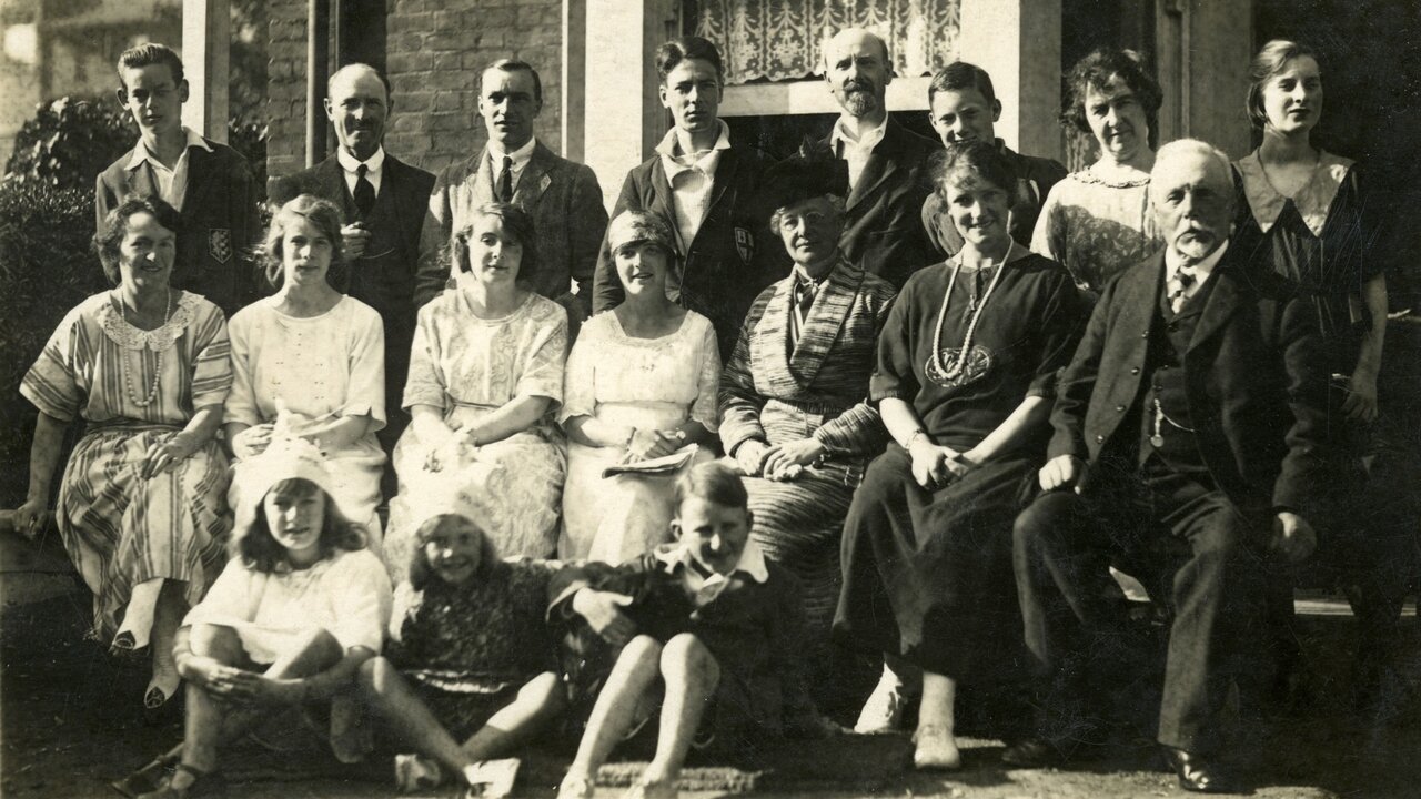 Die Schwarz-Weiß-Fotografie zeigt eine große Gruppe von 18 Personen, die auf einer Veranda sitzen und stehen. Sie sind formell gekleidet und lächeln in die Kamera. Die Szene vermittelt eine familiäre Atmosphäre, wahrscheinlich ein Familientreffen oder eine Feier.