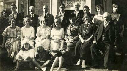 Die Schwarz-Weiß-Fotografie zeigt eine große Gruppe von 18 Personen, die auf einer Veranda sitzen und stehen. Sie sind formell gekleidet und lächeln in die Kamera. Die Szene vermittelt eine familiäre Atmosphäre, wahrscheinlich ein Familientreffen oder eine Feier.