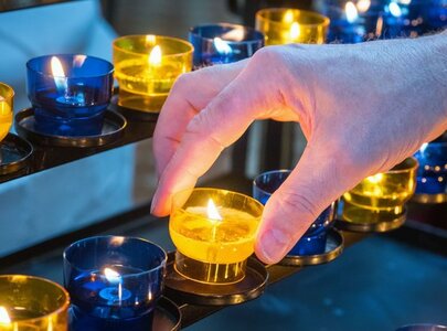 Eine Hand platziert eine brennende gelbe Kerze auf einem Kerzenhalter, der mehrere leuchtende Kerzen in Blau und Gelb zeigt. Die Szene vermittelt eine ruhige und feierliche Atmosphäre.