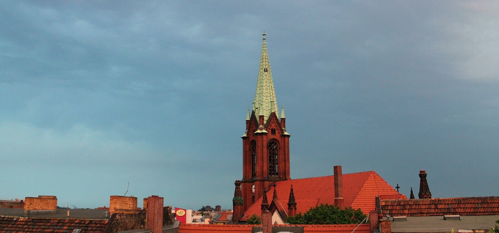 Eine rote Backsteinkirche mit einem hohen, spitzen Turm und einer grünen Kupferhaube ragt über die Dächer der Stadt. Der Himmel ist bewölkt, und die Atmosphäre wirkt ruhig und gedämpft.