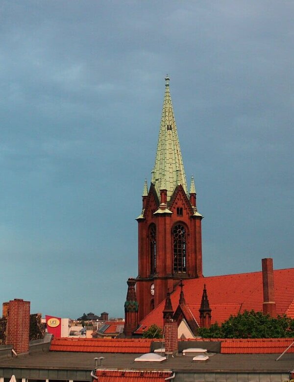 Eine rote Backsteinkirche mit einem hohen, spitzen Turm und einer grünen Kupferhaube ragt über die Dächer der Stadt. Der Himmel ist bewölkt, und die Atmosphäre wirkt ruhig und gedämpft.