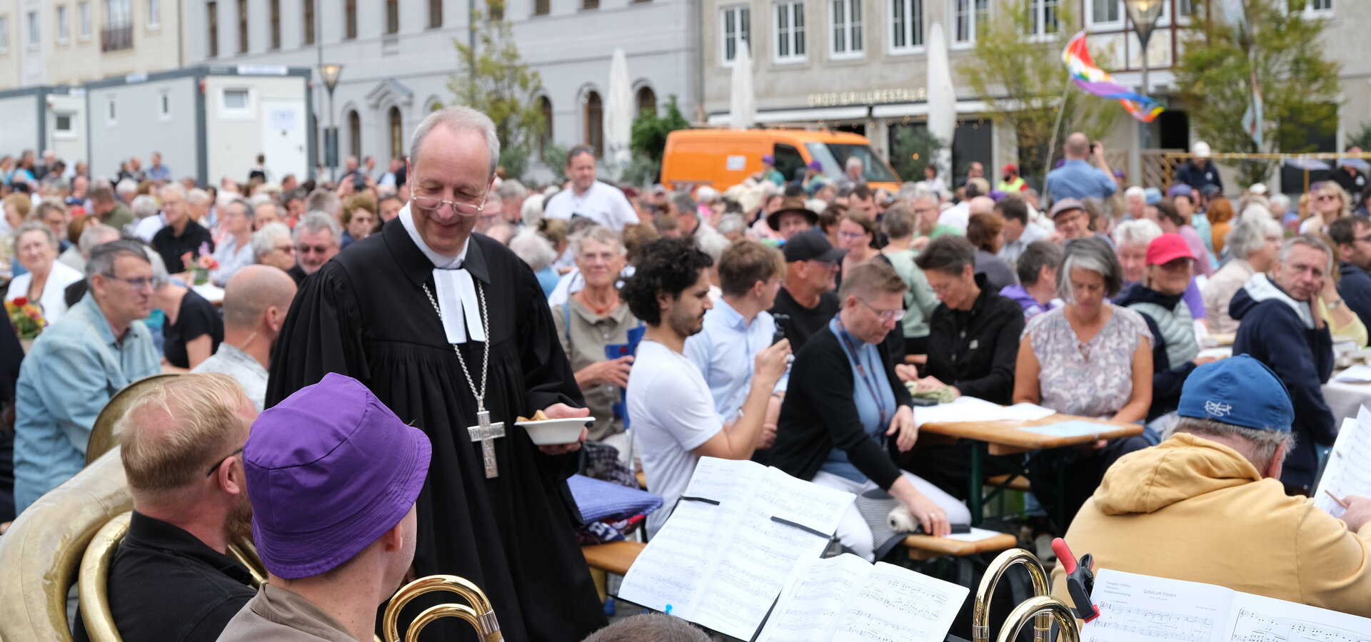 Eine große Menge von Menschen versammelt sich auf einem Platz, während ein Musiker mit einer Tuba im Vordergrund spielt. Ein Mann in einem schwarzen Gewand mit einer weißen Halskrause bewegt sich durch die Menge. Die Atmosphäre wirkt festlich und gesellig.