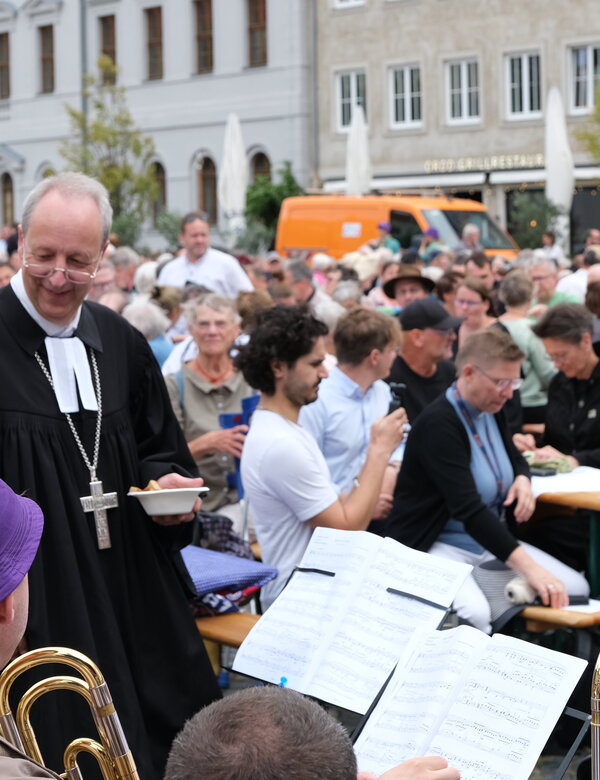 Eine große Menge von Menschen versammelt sich auf einem Platz, während ein Musiker mit einer Tuba im Vordergrund spielt. Ein Mann in einem schwarzen Gewand mit einer weißen Halskrause bewegt sich durch die Menge. Die Atmosphäre wirkt festlich und gesellig.