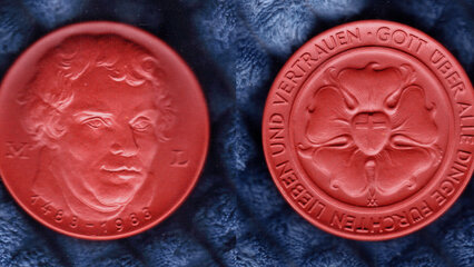 Die Abbildung zeigt eine rote Medaille, auf der eine Darstellung eines Gesichts auf der Vorderseite und ein stilisiertes florales Muster mit dem Text "GOTT ÜBER ALLE DINGEN" auf der Rückseite zu sehen ist. Die Jahreszahlen 1783 und 1983 sind ebenfalls eingraviert.