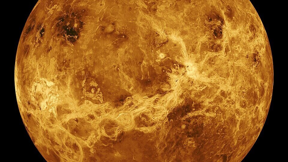 Die Darstellung zeigt den Planeten Venus, der von einer dichten, gelb-orangefarbenen Atmosphäre umgeben ist. Die Oberfläche ist von felsigen und vulkanischen Merkmalen geprägt, die durch Wolken aus Schwefelsäure verborgen sind. Die dunkelsten Stellen deuten auf unterschiedliche geologische Strukturen hin.