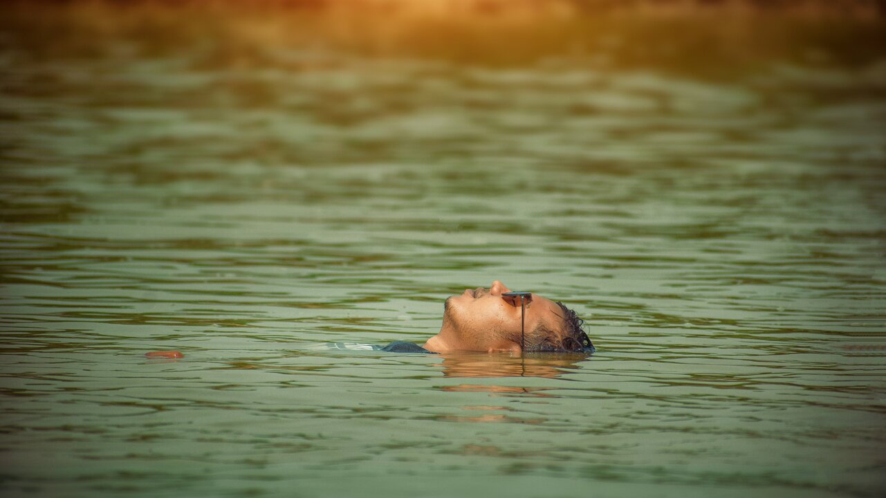 Eine Person liegt auf dem Rücken im Wasser und schaut nach oben. Die Kopfposition ist entspannt, und das Licht reflektiert sanft auf der Wasseroberfläche. Es vermittelt ein Gefühl von Ruhe und Gelassenheit.