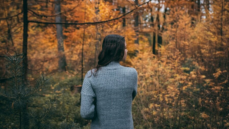 Eine Frau steht mit dem Rücken zur Kamera in einem Wald. Der Herbst hat die Blätter in warmen Orange- und Gelbtönen gefärbt. Die Atmosphäre ist ruhig und natürlich, während die Frau den Anblick der herbstlichen Landschaft genießt.