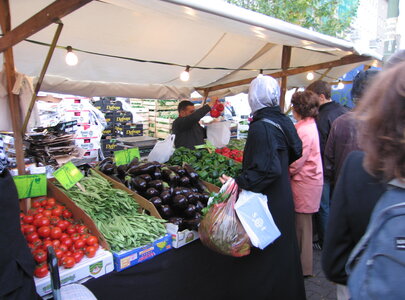 Ein Marktstand mit frischem Gemüse: rote Tomaten, grüne Bohnen, Auberginen und Kräuter sind gut sichtbar. Menschen stehen in einer Schlange, während ein Verkäufer eine Tüte mit Gemüse überreicht. Ein Zelt bietet Schutz vor der Witterung.