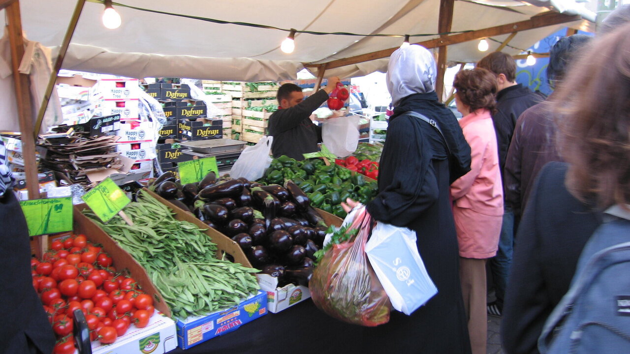 Ein Marktstand mit frischem Gemüse: rote Tomaten, grüne Bohnen, Auberginen und Kräuter sind gut sichtbar. Menschen stehen in einer Schlange, während ein Verkäufer eine Tüte mit Gemüse überreicht. Ein Zelt bietet Schutz vor der Witterung.